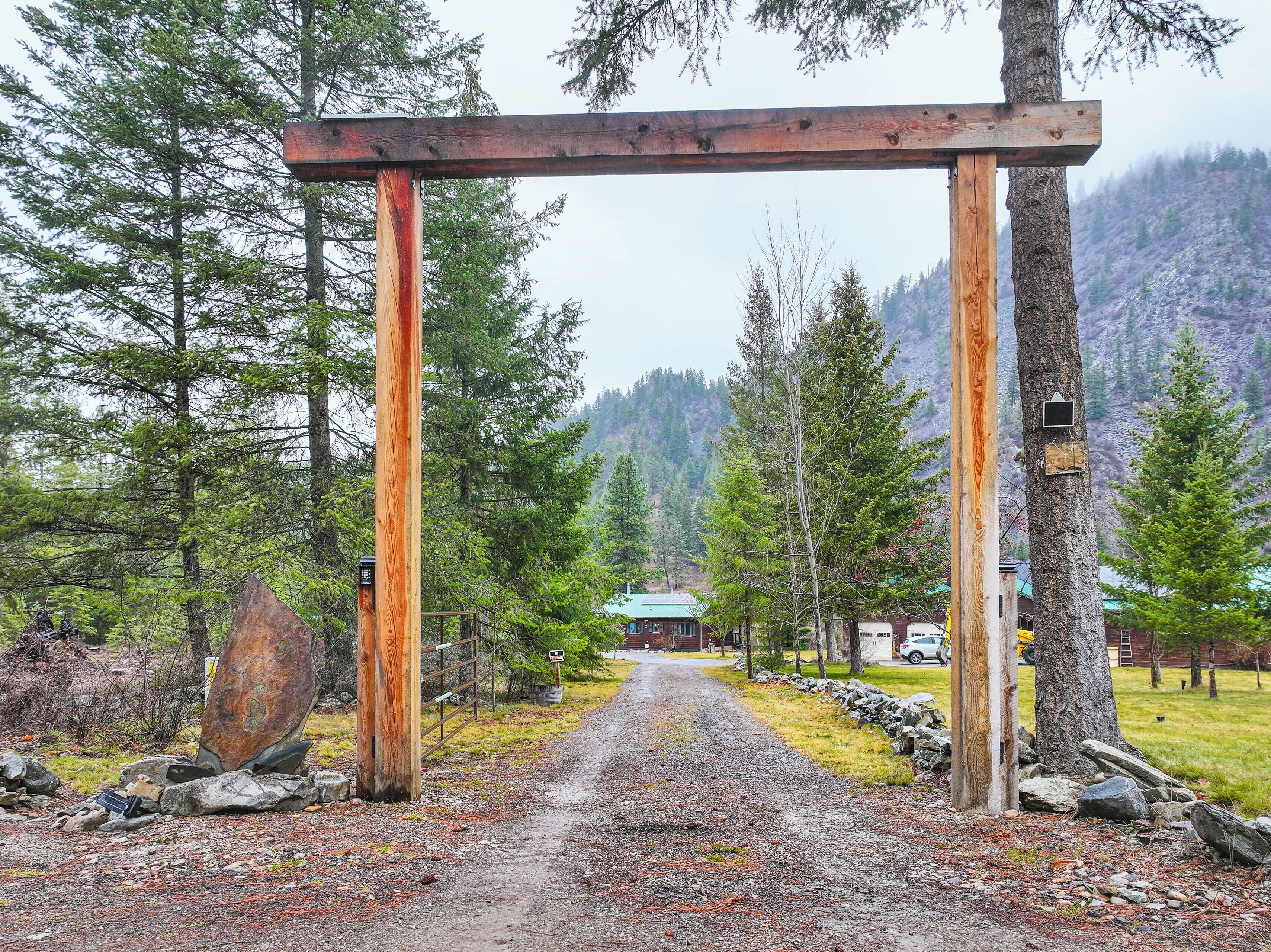 A rustic wooden archway leading to a building with a green roof, surrounded by trees and a mountainous backdrop.