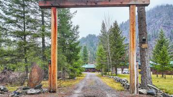 A rustic wooden archway leading to a building with a green roof, surrounded by trees and a mountainous backdrop.