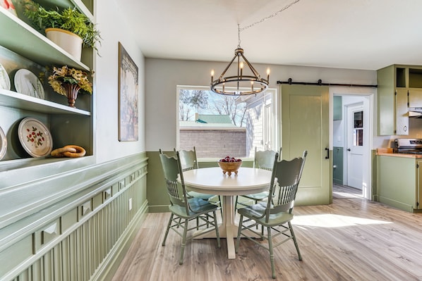 A dining area with a round table, four chairs, and a chandelier. There is a window with a view of a house and trees.