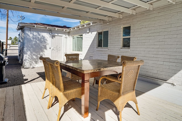 A covered patio with a wooden table and wicker chairs, white brick walls, and a small shed in the background.
