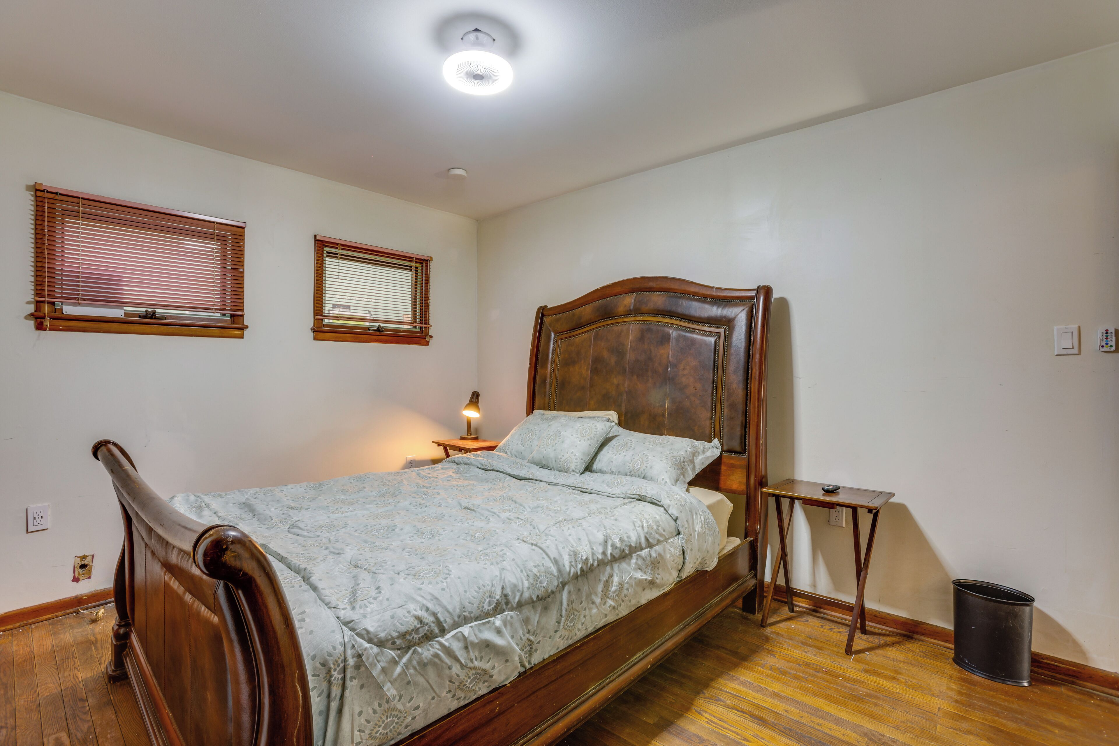 A bedroom with a wooden bed, two windows with blinds, a bedside table, and a trash can.