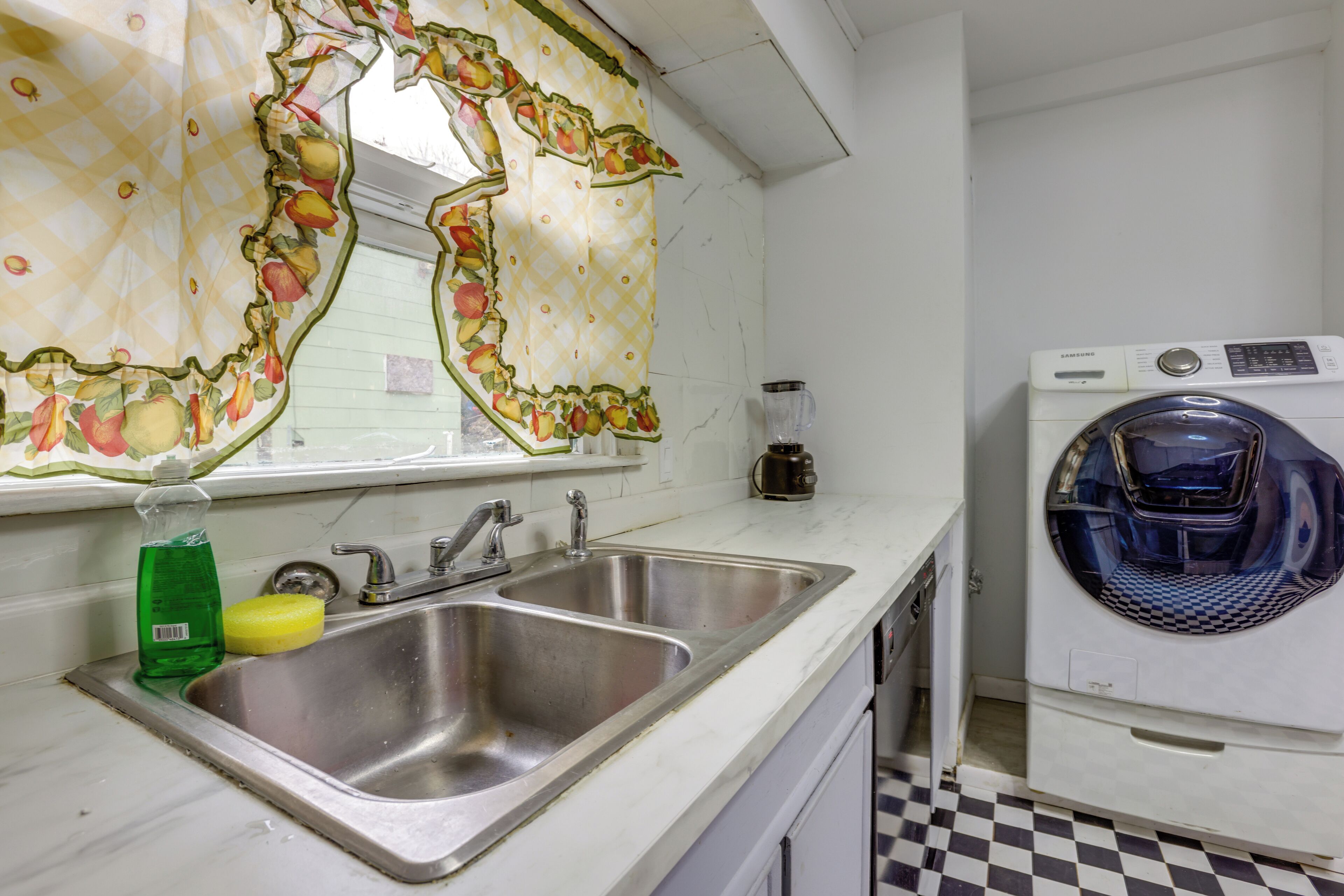 A kitchen with a double sink, a washing machine, and a window with a fruit-patterned curtain.
