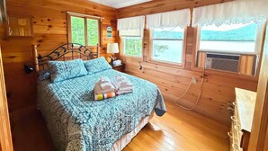 A wooden cabin bedroom with a bed, a nightstand, a lamp, and windows with curtains.
