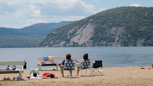 A beach with people relaxing on chairs and a table, overlooking a lake with hills in the background.