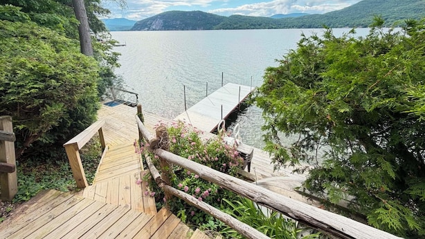 A wooden deck leading to a dock over water, surrounded by greenery and hills in the background.