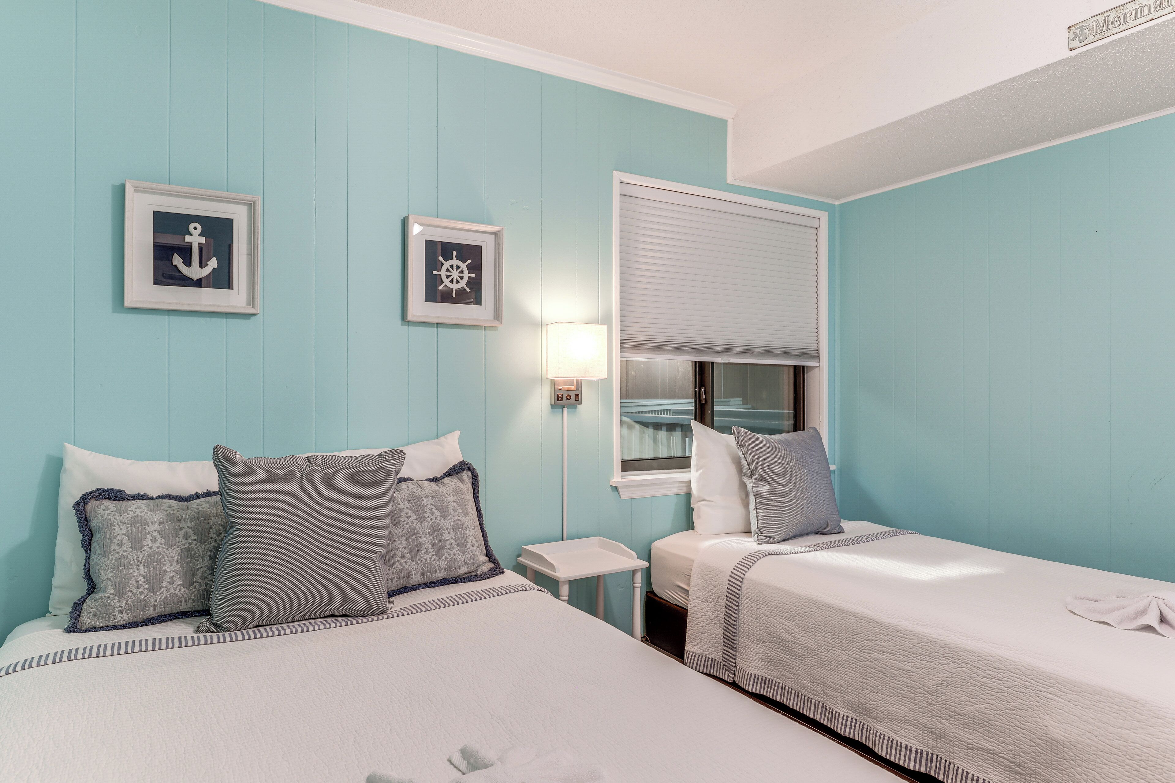 A room with two beds, a window with blinds, a wall-mounted lamp, and two framed pictures.