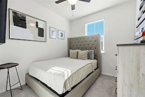 Une chambre à coucher avec une tête de lit capitonnée, un lit recouvert d’une couette blanche, un petit tabouret, un ventilateur de plafond et une fenêtre avec des stores.
