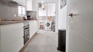 Ett modernt kök med vita skåp, en bänkskiva i granit, en inbyggd ugn och en mikrovågsugn. Till höger finns en svart soptunna, och en dörr leder till ett tvättrum med tvättmaskin och torktumlare.