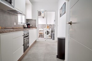 A modern kitchen with white cabinets, a granite countertop, a built-in oven, and a microwave. There is a black trash bin to the right and a door leading to a laundry area with a washing machine and dryer. - Tucuman 3 Los Huertos Nerja by Casasol (Nerja)