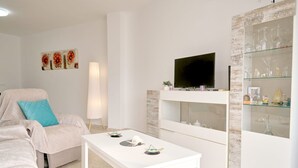 Ett vardagsrum med en vit soffa, ett soffbord, en tv som är monterad på ett vitt skåp och ett vitrinskåp med dekorativa föremål.