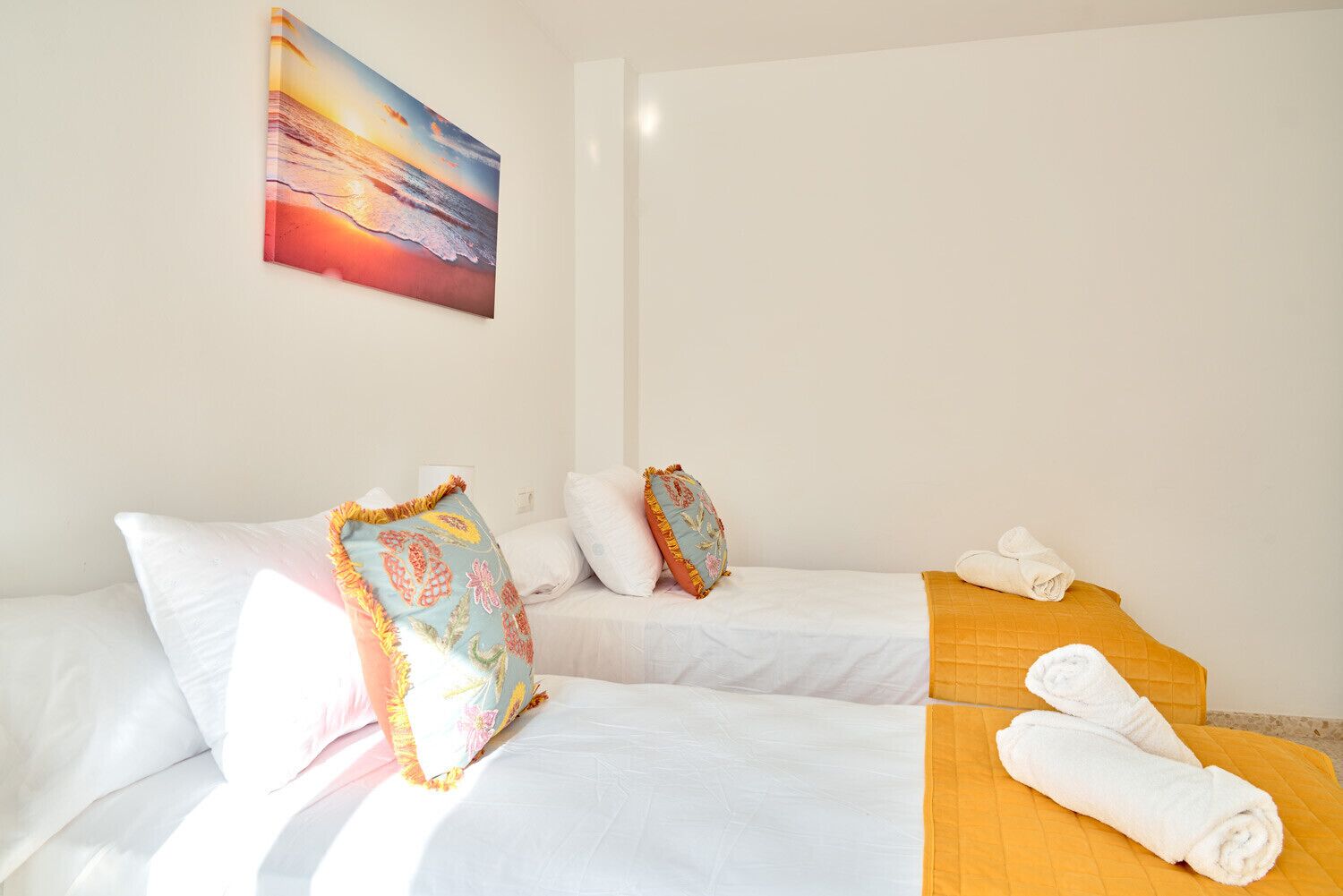 A hotel room with two beds, a painting of a sunset, and a yellow bed runner.