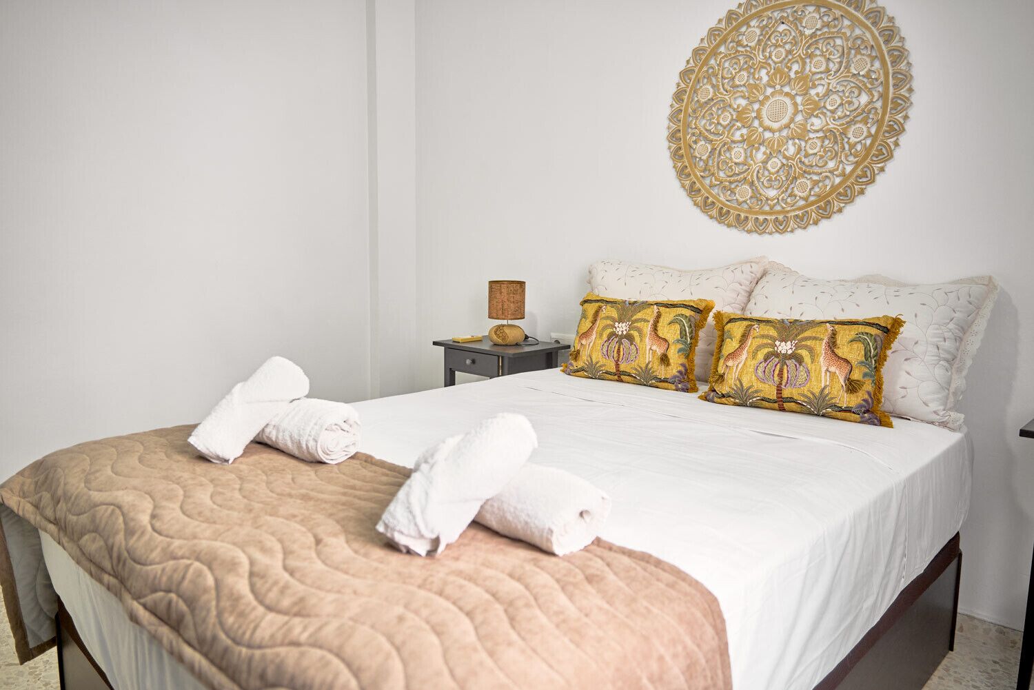 A neatly made bed with a beige quilt, two white towels, and two decorative pillows with animal patterns.