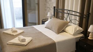 Una cama bien hecha con una colcha color beige y almohadas, dos toallas enrolladas sobre la cama, una mesita de noche con lámpara, y vistas a un balcón a través de una puerta de cristal.