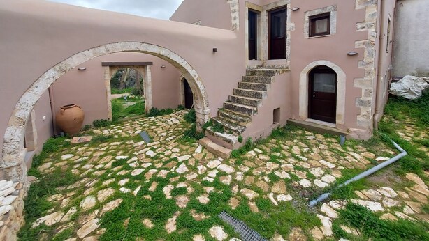 A building with a stone staircase leading to a door, a large archway, and a small courtyard with a stone floor and greenery. - Conastasia - Concona Houses (Rethymno)