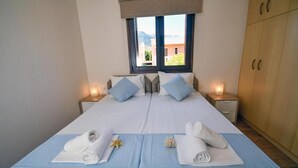 A double bed with a blue bedspread, two nightstands with lamps, a window with a view of buildings and mountains, and a wooden wardrobe. - Pietra Sole Villa (Makry Gialos)