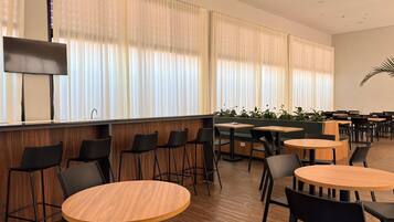A modern indoor dining area with round wooden tables and black chairs, large windows with white curtains, and a wooden wall paneling.