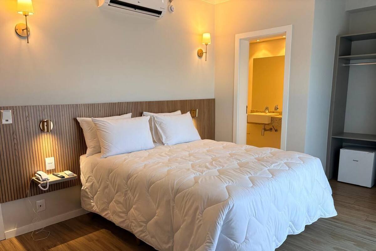 Ein Hotelzimmer mit einem großen Bett, Nachttischlampen, einem kleinen Kühlschrank und einem durchgegangene Tür erkennbaren Badezimmer.