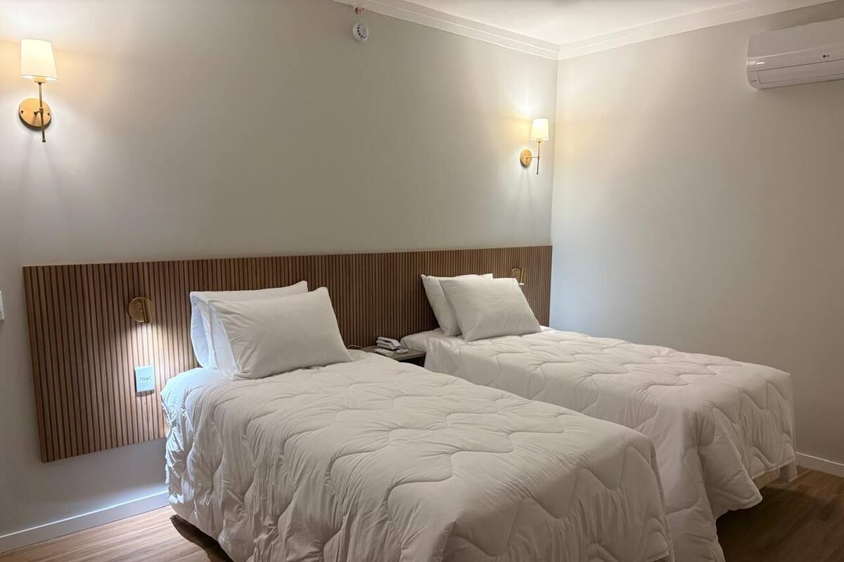 Ein Hotelzimmer mit zwei Betten, einem Holzkopfstück, Wandleuchten und einer fest installierten Klimaanlage.