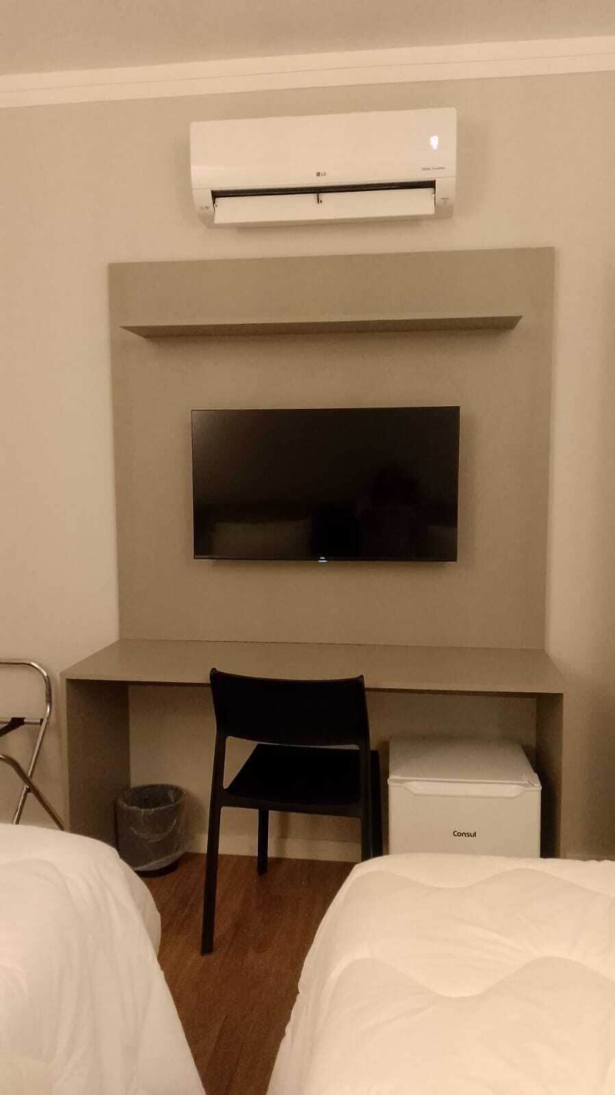 Hotelkamer met een bed, een bureau met stoel, een televisie, een airconditioner en een klein wit kastje.