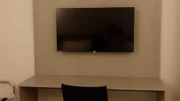 Hotelkamer met een bed, een bureau met stoel, een televisie, een airconditioner en een klein wit kastje.