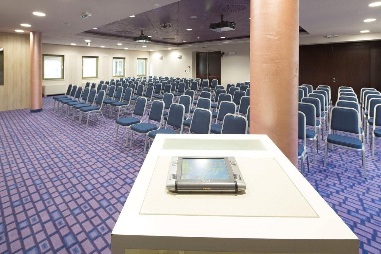 A conference room with rows of blue chairs, a white table with a laptop, and a patterned carpet.