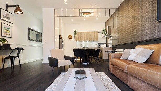 A modern living room with a brown leather sofa, a dining area with a wooden table and chairs, and a kitchen in the background. - La Suite Urbaine - Place d'Erlon - Bulle de Calme (Reims)