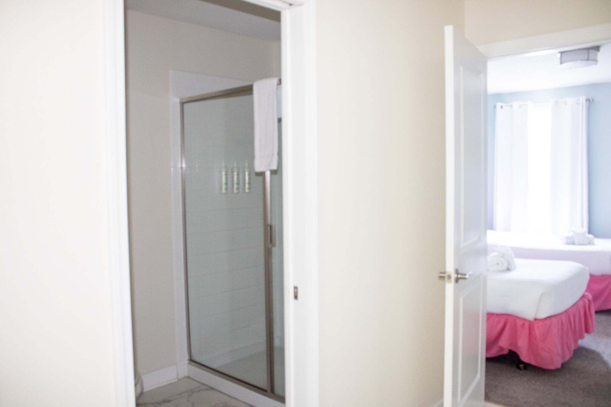 A room with a glass shower door, a bed with a white and pink bedspread, and a window with curtains.
