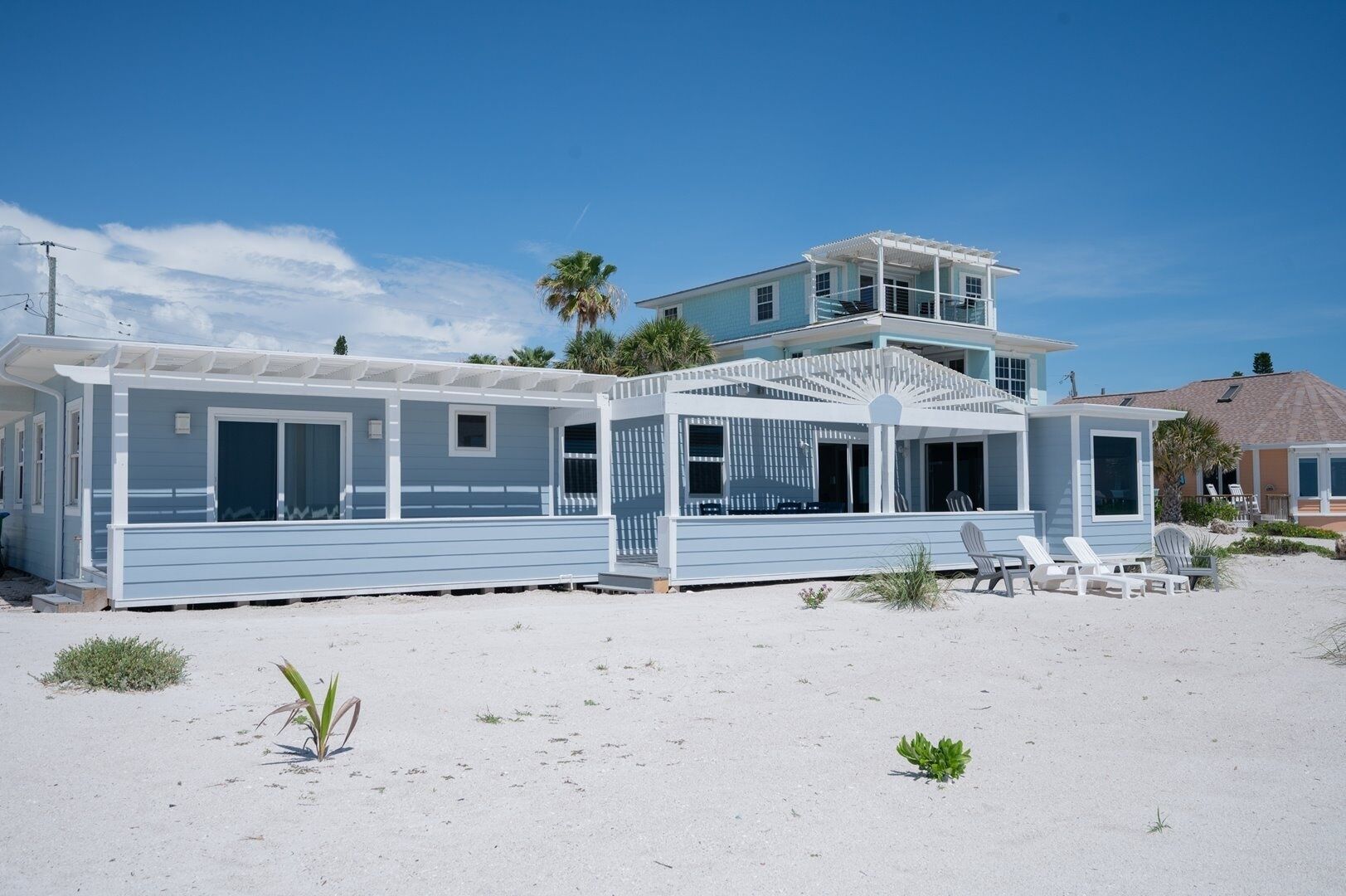 A beachfront house with a two-story structure, a covered porch, and a sandy beach area with lounge chairs.