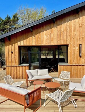 Una construcción de madera con terraza, asientos al aire libre y ventanas amplias.