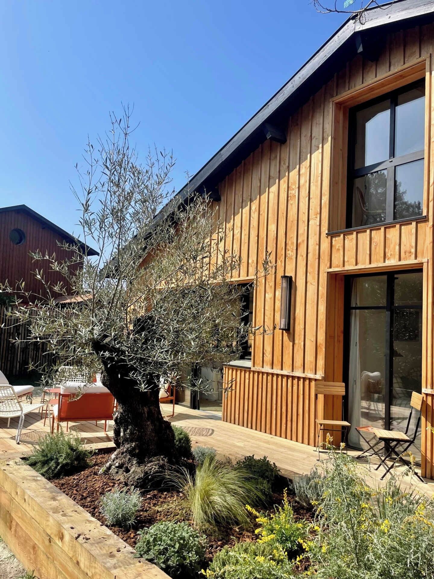 Un bâtiment en bois doté d’une grande fenêtre et d’un patio aménagé avec du mobilier d’extérieur.