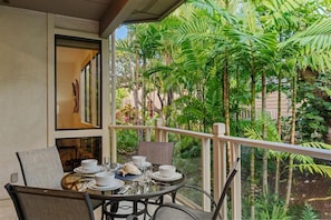 A balcony with a table set for dining, surrounded by lush tropical plants. - Grand Champions 185 2 Bedroom Condo (Kihei)