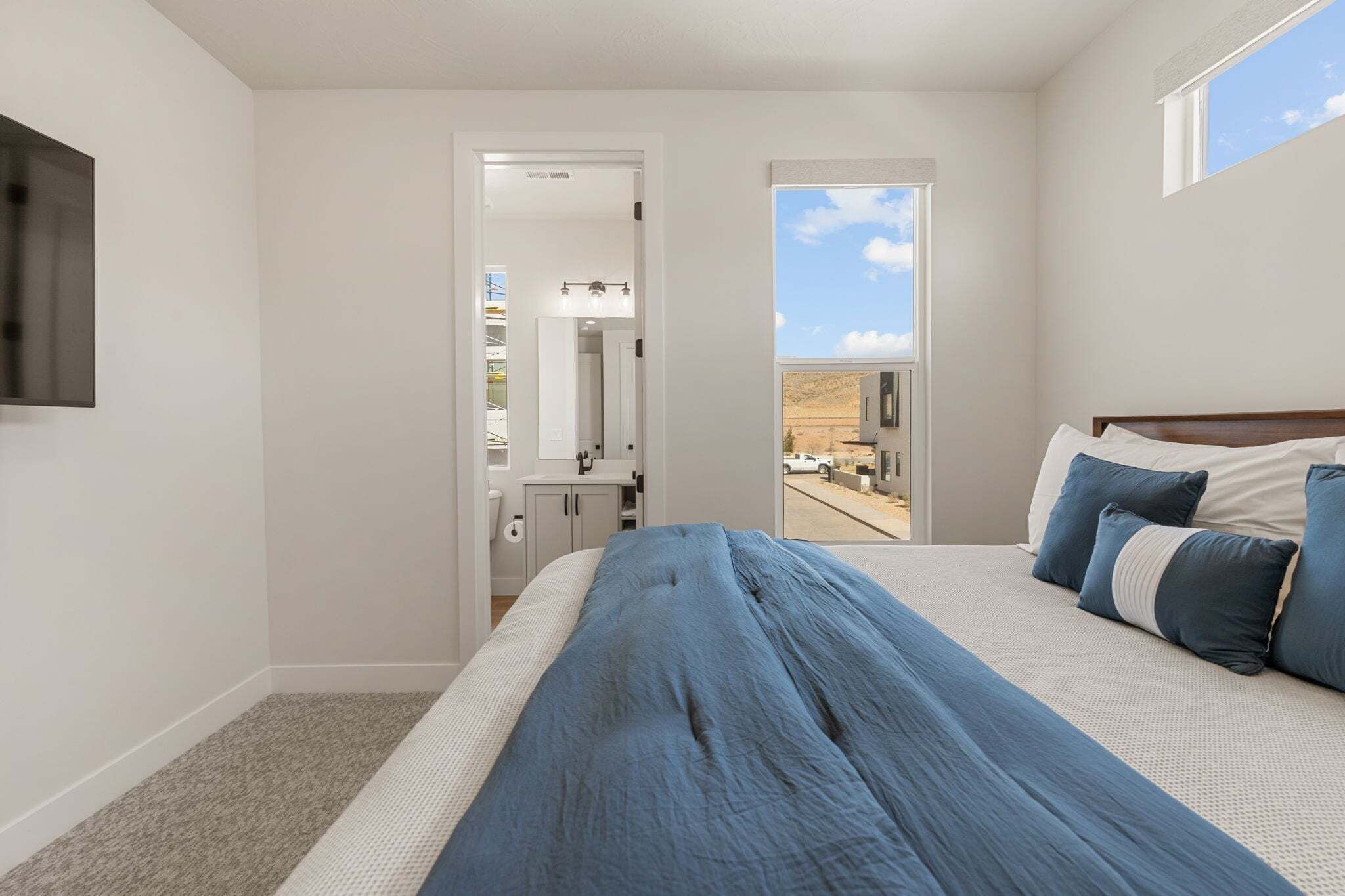 A bedroom with a large bed, a TV mounted on the wall, and a view of a desert landscape through a window.