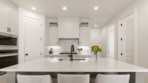 A modern kitchen with white cabinetry, a central island with a sink, and three bar-style chairs. - Rockaction Destination 5 Bedroom Townhouse (St. George)