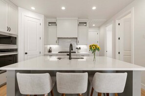 A modern kitchen with white cabinetry, a central island with a sink, and three bar-style chairs. - Rockaction Destination 5 Bedroom Townhouse (St. George)