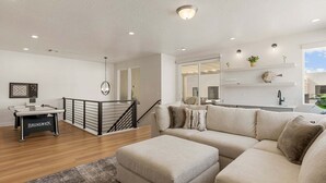 A modern living room with a beige sofa, a coffee table, and a pool table. There is a staircase with a black railing leading to a balcony. - Rockaction Destination 5 Bedroom Townhouse (St. George)