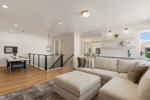 A modern living room with a beige sofa, a coffee table, and a pool table. There is a staircase with a black railing leading to a balcony. - Rockaction Destination 5 Bedroom Townhouse (St. George)