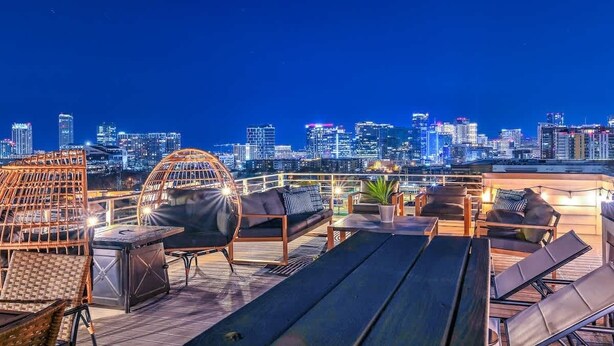 Eine Dachterrasse mit Liegestühlen, einem Tisch und einem Korbsessel mit Blick auf eine beleuchtete Stadtlandschaft in der Nacht.