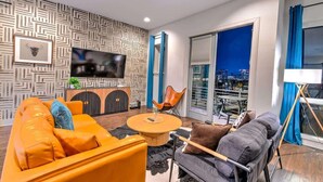 Ein modernes Wohnzimmer mit Sofa, Sessel und Couchtisch, einem Flachbildfernseher und Blick auf die Stadt durch ein großes Fenster.