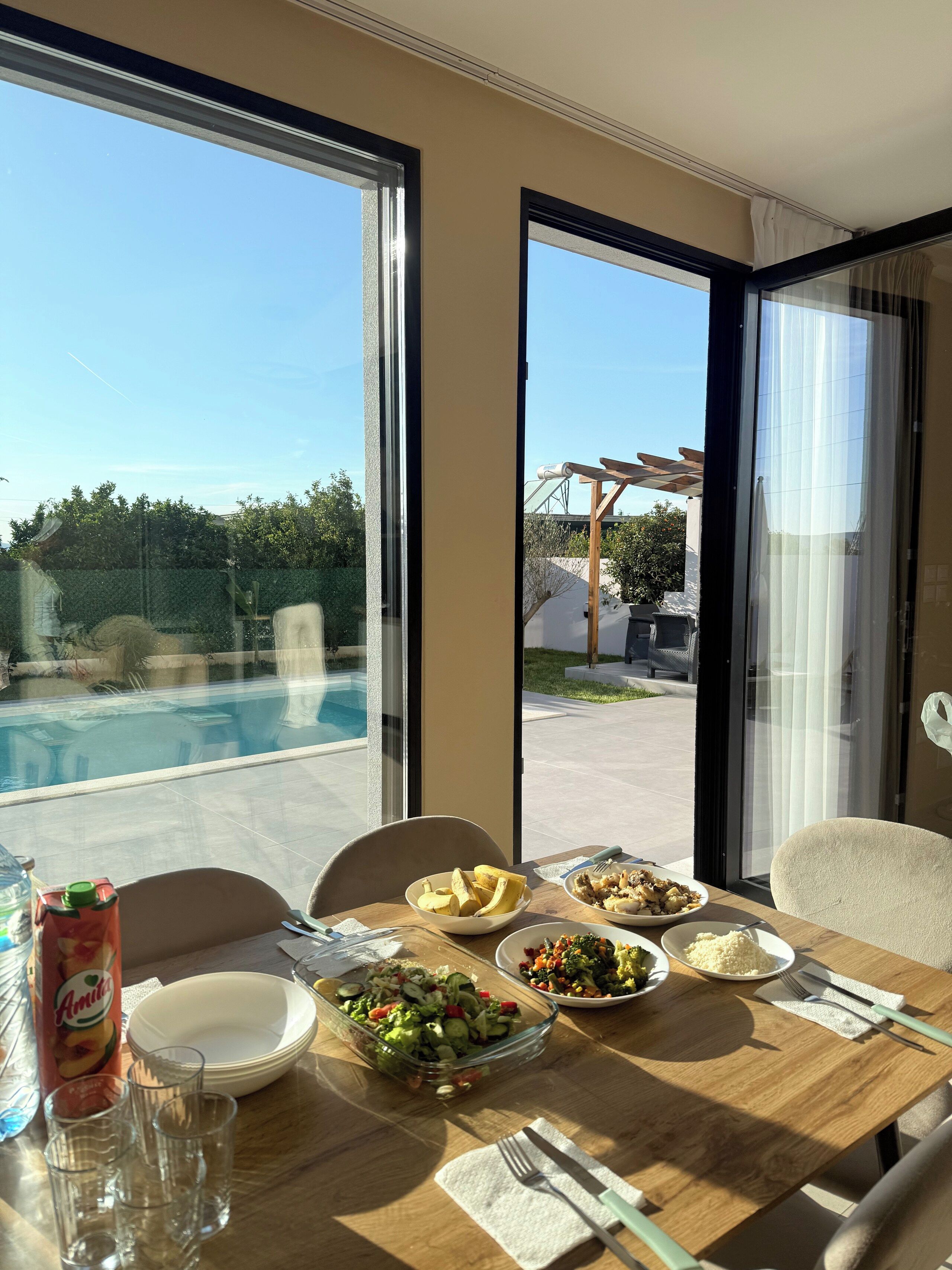 Ein Esstisch mit verschiedenen Speisen, einer Flasche und Gläsern, vor einer Pool- und Außenbereichsitzgelegenheit.