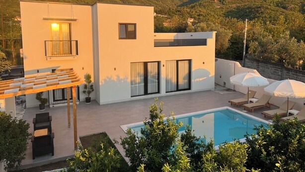 Ein modernes, zweistöckiges Haus mit Swimmingpool, Sitzgelegenheit im Freien und einer Bergkulisse im Hintergrund.
