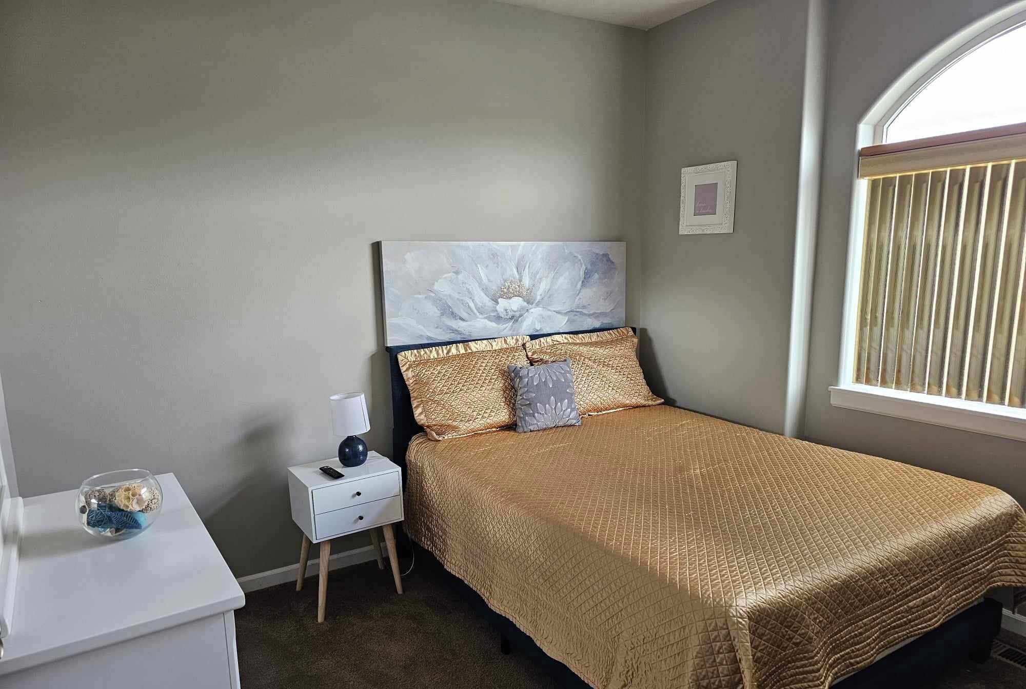 A bedroom with a bed, a nightstand, a lamp, a decorative vase, and a window with blinds.