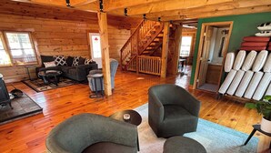A cozy cabin interior with a wooden floor, a sofa, two armchairs, a small table, and a staircase leading to an upper level.