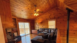 A cozy cabin interior with a wooden ceiling, a sofa set, a coffee table, a fireplace, and a sliding glass door leading to a deck.