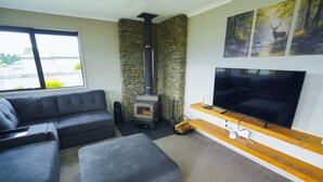 A living room with a stone fireplace, grey sofas, a flat-screen TV, and a wooden shelf. - Southern Peaks by Aotearoa Escapes (te anau)