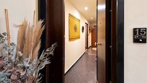 Un pasillo con una puerta marcada con el número 15, un tapiz decorativo y una planta con plumas de plumas y hojas de eucalipto.