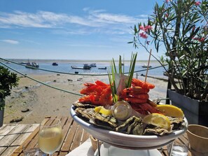 Eine Platte mit Meeresfrüchten, Austern, Garnelen und Zitronenscheiben auf einem Tisch am Strand, im Hintergrund sind Boote zu sehen.