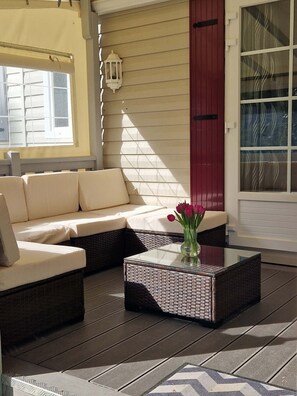 A sunlit patio with a wicker sofa, a glass coffee table with a vase of flowers, and a red door. - Nell's Cabin (Lège-Cap-Ferret)