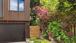 Una casa moderna con un amplio portón de garaje, cerca viva de madera y un jardín con diversas plantas y una enredadera.