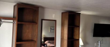 A hotel room with a bed, a wooden wardrobe, a mirror, a television, and a small table with a lamp.