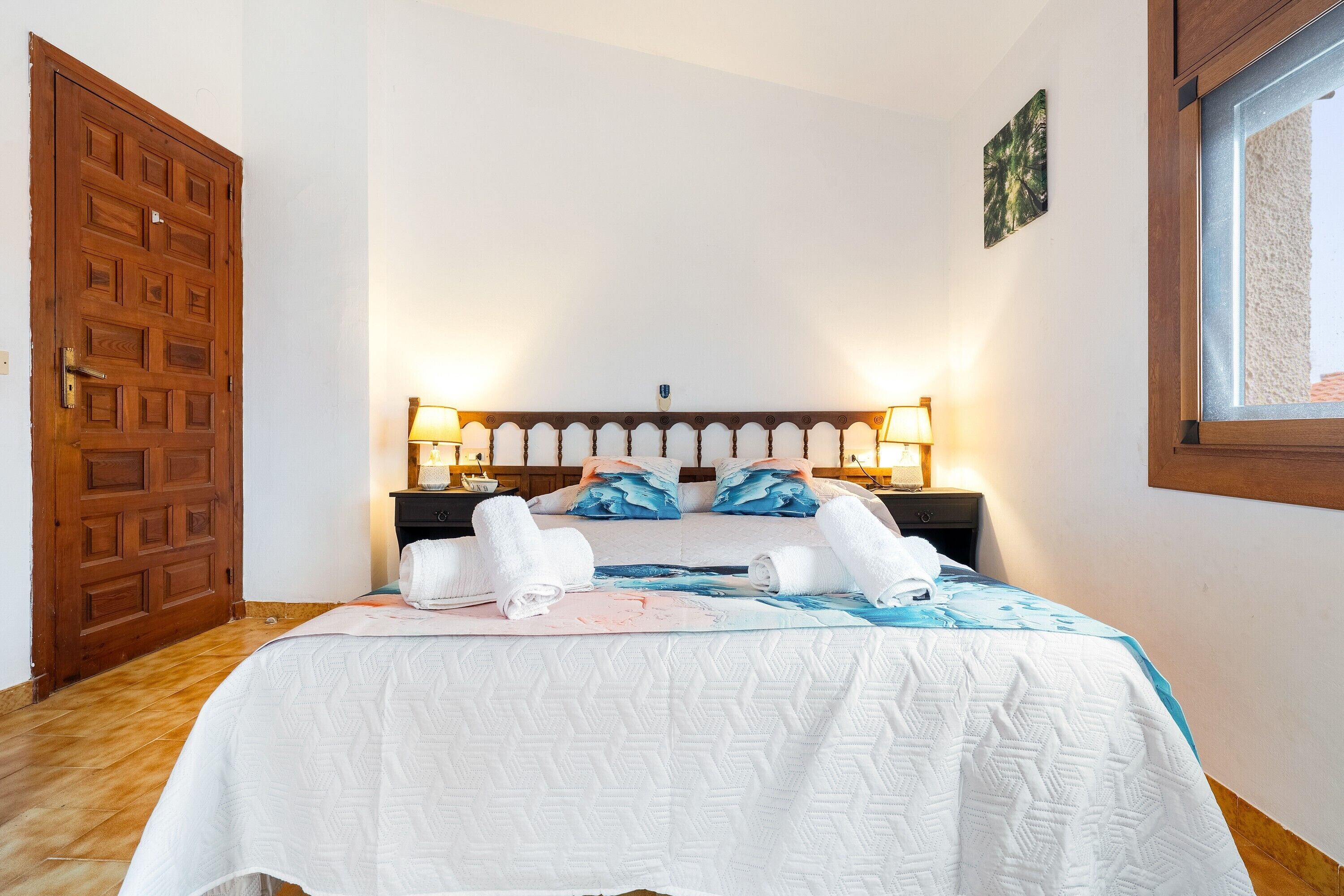 A bedroom with a wooden door, a bed with a white quilt, two bedside lamps, and a window.